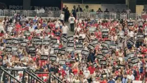 Stadium crowd stands, raising black “Turning Point” signs; red-white-blue outfits fill the seats during the Charlie Kirk memorial in Glendale, Arizona.