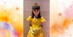 Young girl wearing a yellow princess dress with a tiara and gloves, smiling outdoors in front of a garage door.