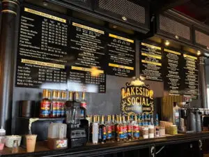 The ordering counter at Lakeside Social coffee shop, showing large black and gold menu boards filled with drink and food options hanging above a counter lined with flavoring syrups and coffee equipment.