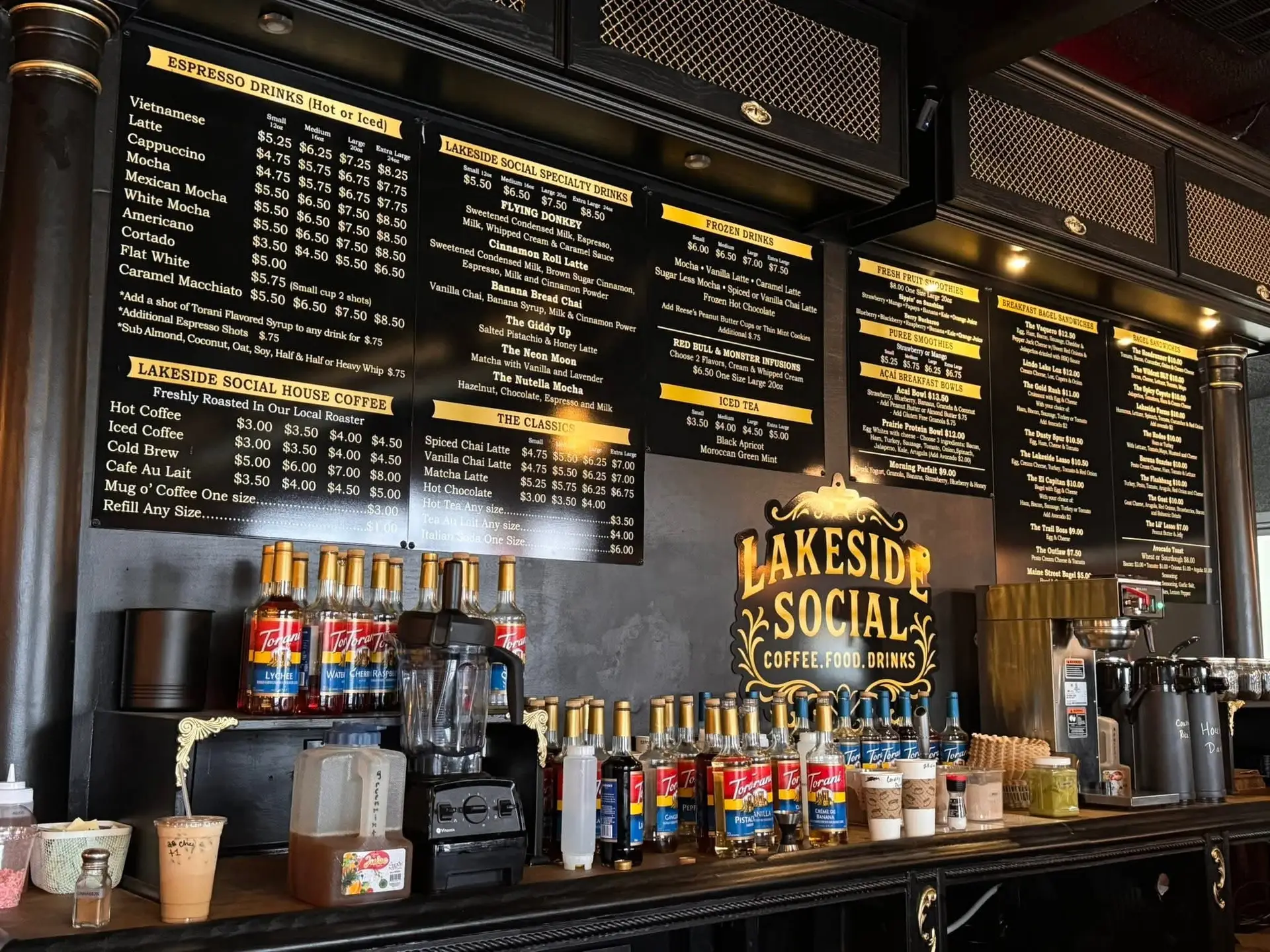 The ordering counter at Lakeside Social coffee shop, showing large black and gold menu boards filled with drink and food options hanging above a counter lined with flavoring syrups and coffee equipment.